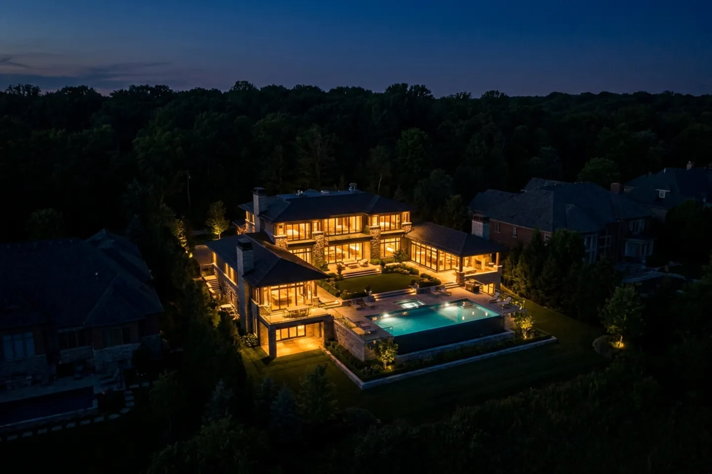 An aerial view at night of one large luxury estate fully lit across its facade, pool, and grounds, surrounded by darkened neighboring properties