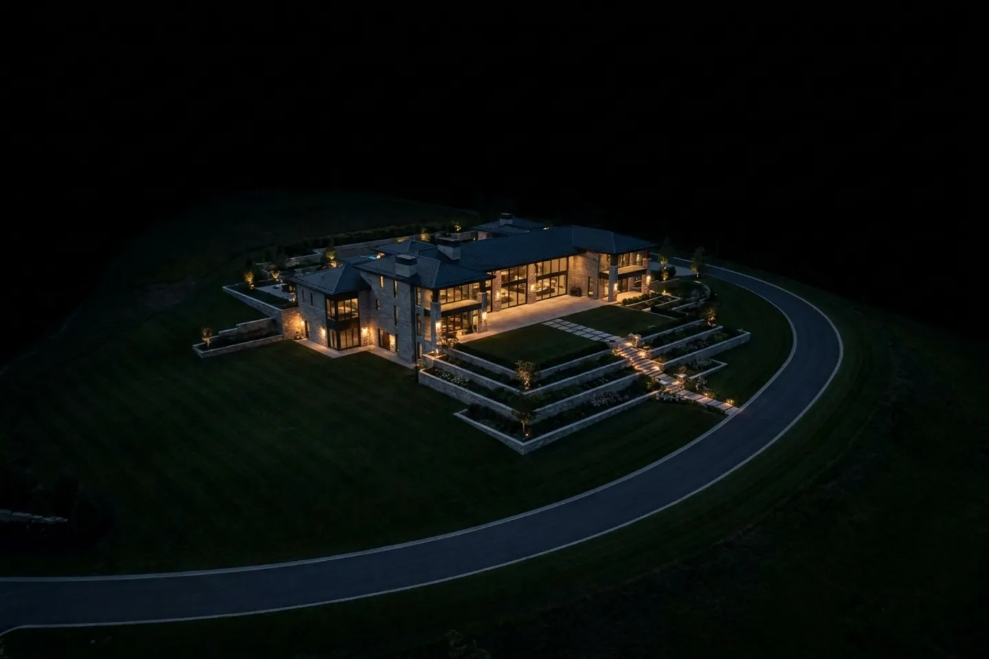 An aerial view at night of an isolated ultra-luxury estate on a large private hillside lot, its lights the only warmth against dark grounds